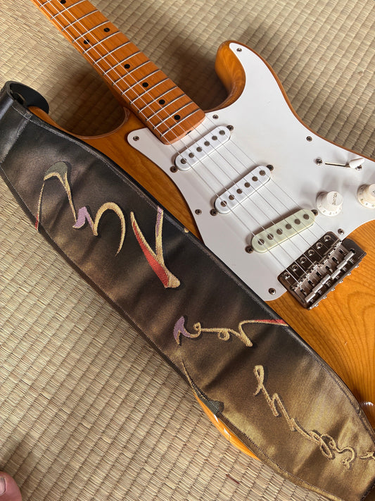 OGF wide guitar strap with a dark woven pattern laid across a natural-finish electric guitar on tatami.